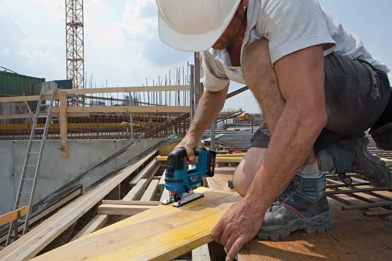 Un trabajador que usa equipo de seguridad corta una tabla de madera con una sierra caladora inalámbrica en un sitio de construcción.