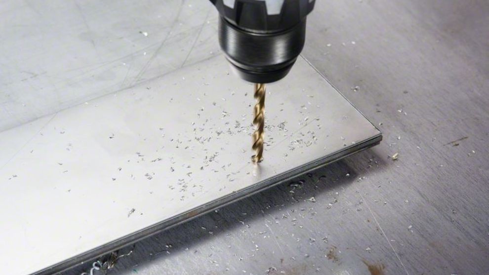 Uma broca penetra em uma chapa de metal brilhante com aparas espalhadas ao redor.