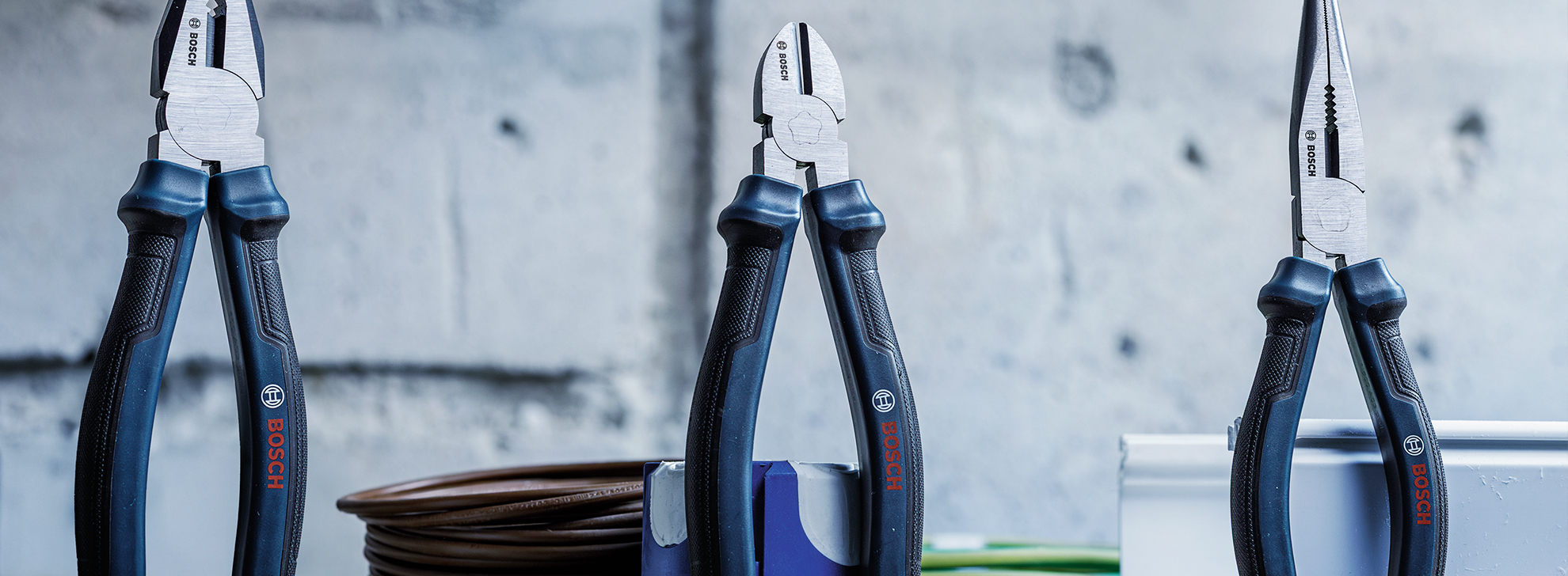 Três alicates profissionais Bosch estão exibidos em uma superfície de trabalho contra uma parede cinza texturizada. Os alicates possuem cabos de borracha preta e mandíbulas de metal brilhante, dispostos com designs variados: um para agarrar, um para cortar e um para tarefas de ponta fina. Perto, uma bobina de fio elétrico marrom e algumas ferramentas coloridas estão parcialmente visíveis. A atmosfera é industrial e funcional, típica de um ambiente de oficina.