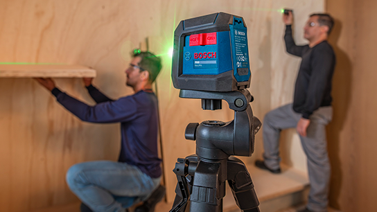 Um nível a laser Bosch Professional em um tripé, com dois homens ajustando prateleiras em uma parede de madeira.