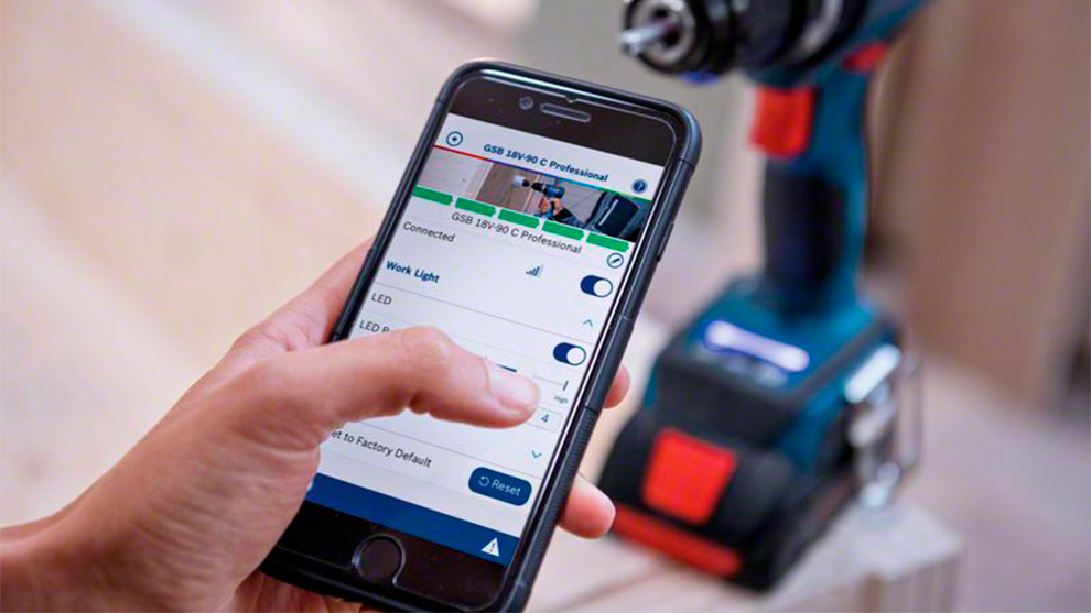 Uma mão segura um smartphone mostrando as configurações da ferramenta Bosch ao lado de uma furadeira.
