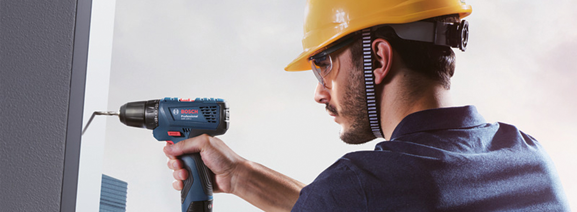 Um homem com um capacete de segurança amarelo usa uma furadeira Bosch Professional em uma parede.