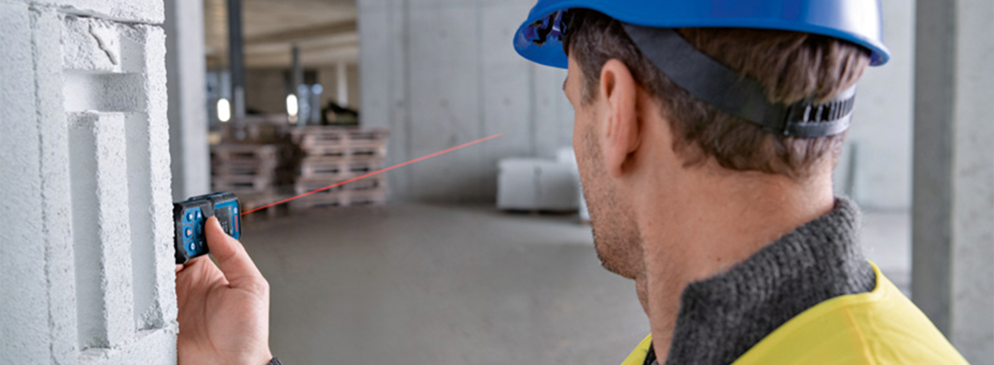 Um homem com um capacete azul usa um medidor de distância a laser Bosch em um armazém.