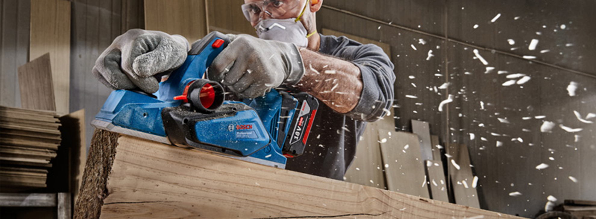 Uma pessoa usa um plaina azul Bosch Professional em uma tábua de madeira, lascas voando.