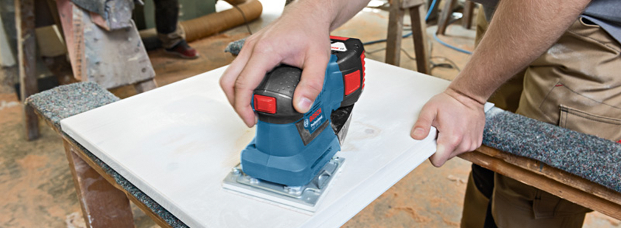 Uma lixadeira Bosch Professional é utilizada em uma peça de trabalho branca sobre uma mesa.