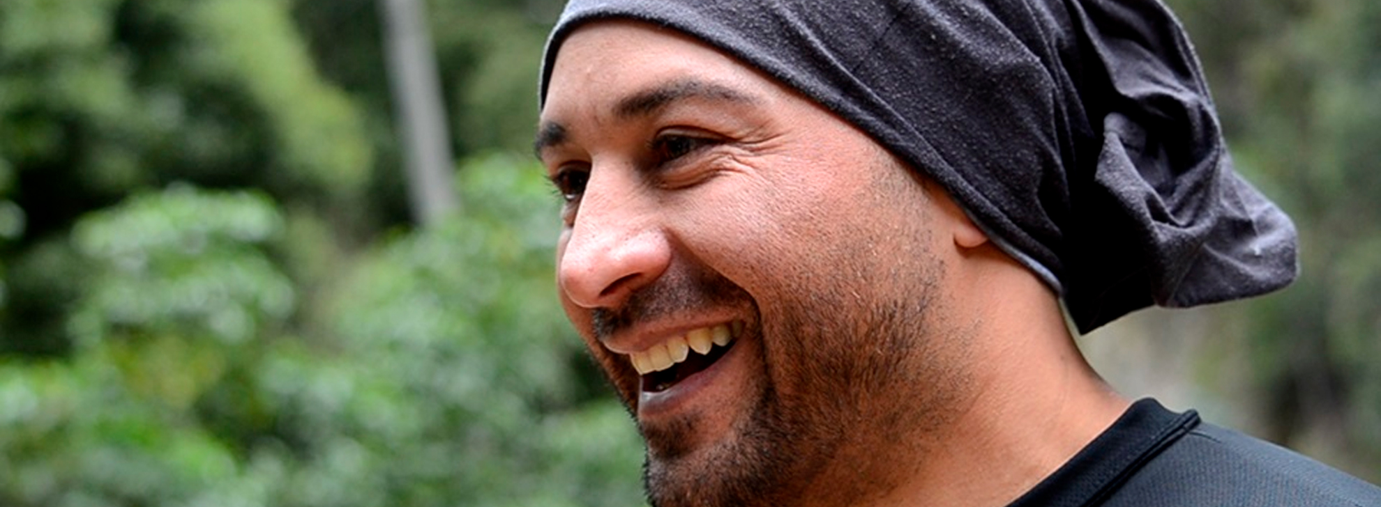 Um homem com um lenço escuro sorri em um ambiente de floresta verde.