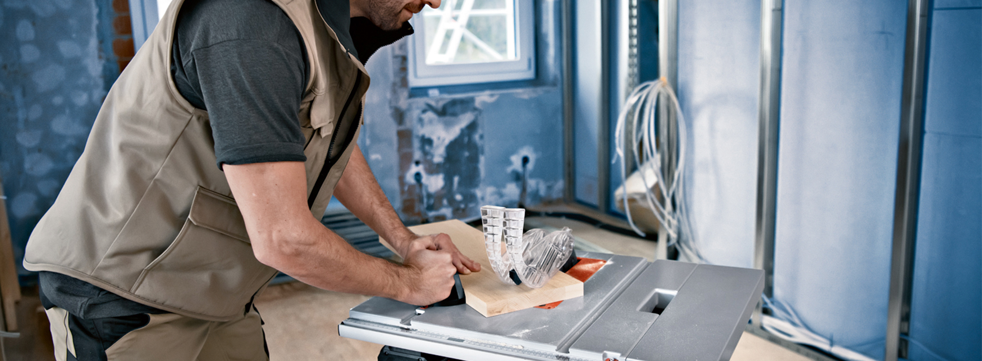 Um homem em um colete trabalha em uma serra de mesa em uma sala em construção.