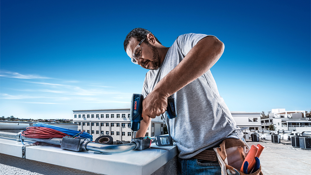 Um trabalhador usando uma furadeira Bosch Professional em um telhado sob um céu azul.