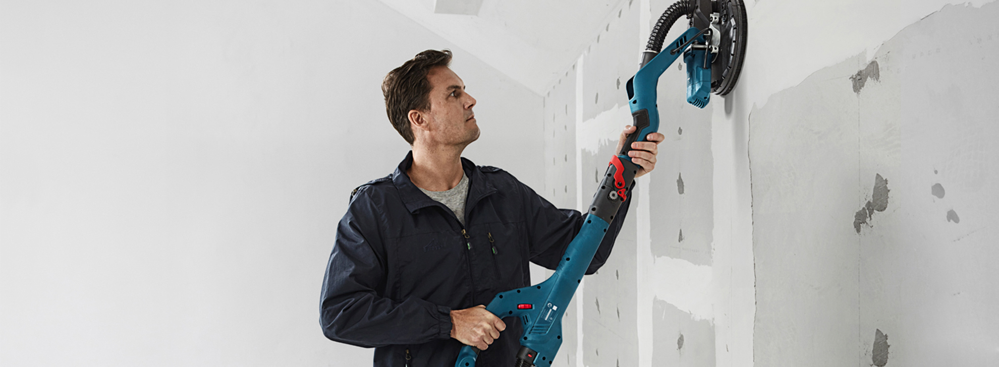 Um homem usa uma lixadeira de drywall Bosch Professional em uma parede branca.