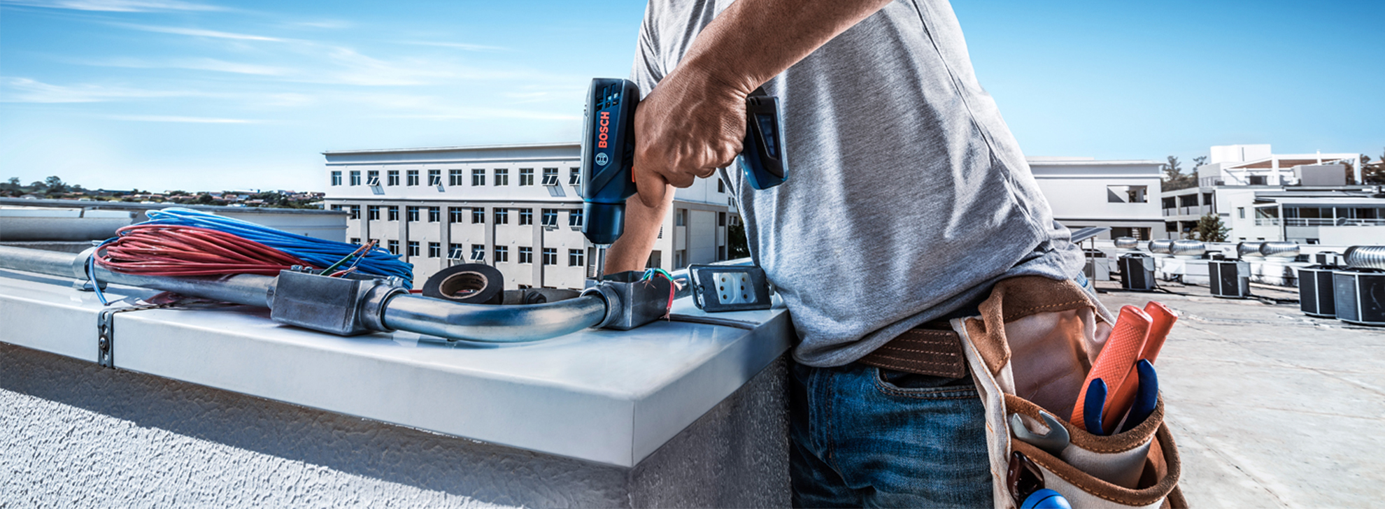 Um trabalhador segura uma ferramenta azul da Bosch perto de um telhado com cabos e edifícios.
