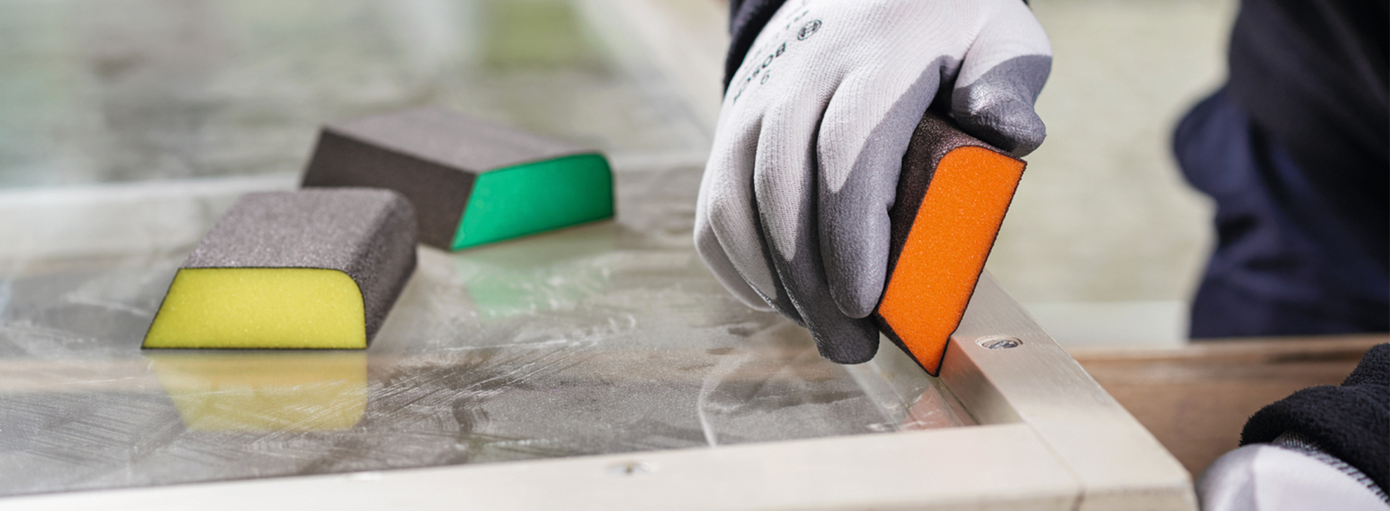 Um trabalhador com luvas usa uma ferramenta laranja em uma superfície com esponjas próximas.