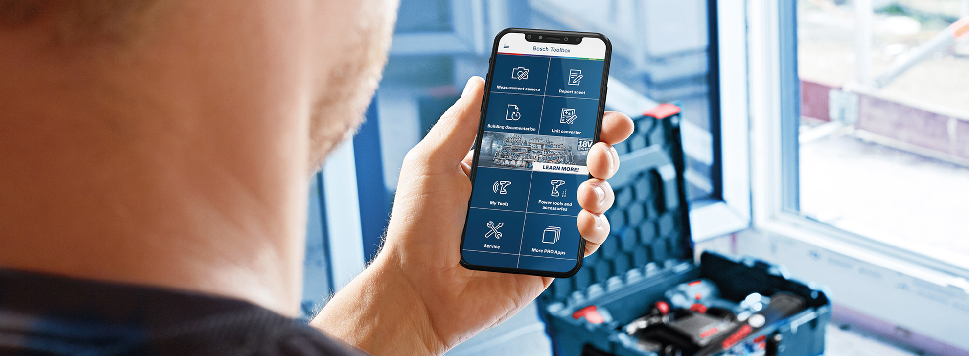 Uma pessoa segura um smartphone exibindo o aplicativo Bosch Toolbox; caixa de ferramentas próxima.