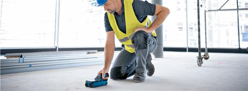 Um homem está de joelhos no chão, usando uma ferramenta azul da Bosch, vestindo um colete amarelo.