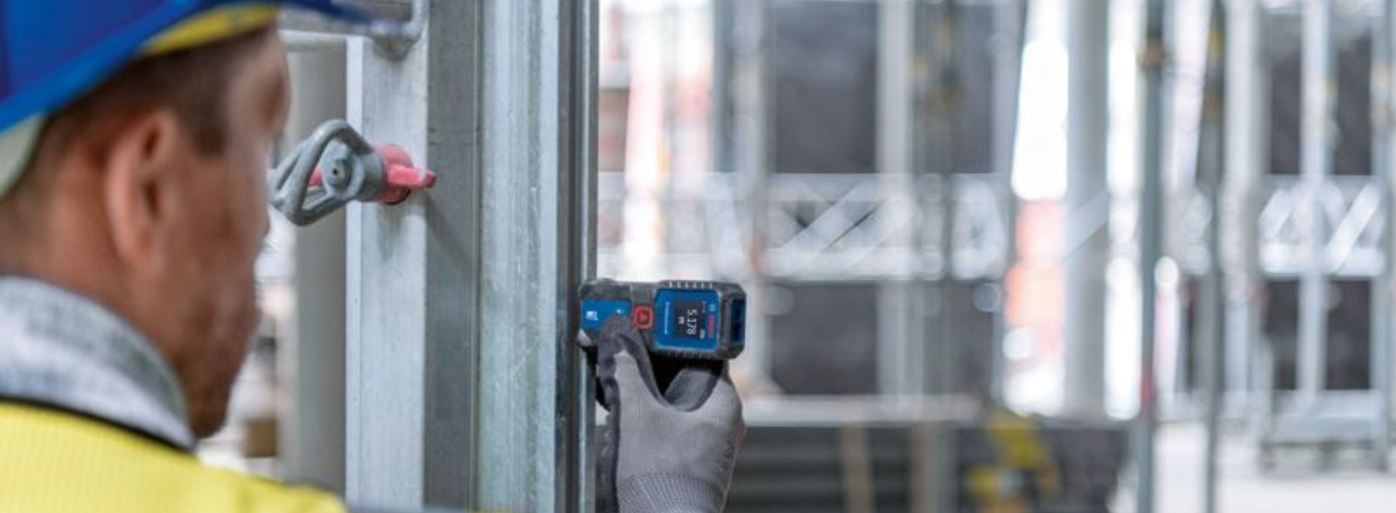 Um trabalhador mede uma viga de aço com uma ferramenta azul da Bosch em um canteiro de obras.