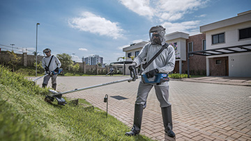 Dois trabalhadores com equipamentos de proteção cortam grama perto de casas modernas.