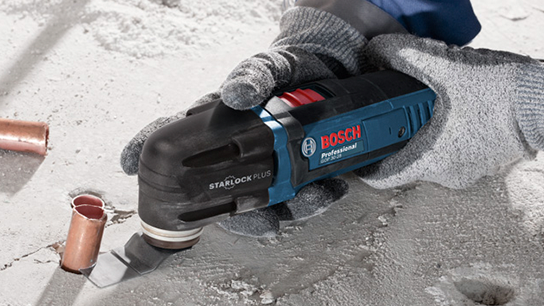 Uma ferramenta oscilante Bosch Professional STARLOCK PLUS está sendo usada para cortar uma superfície de concreto, com tubos de cobre próximos. A ferramenta tem um design preto e azul, e o usuário está usando luvas cinzas, mostrando atividade focada em um ambiente de construção.