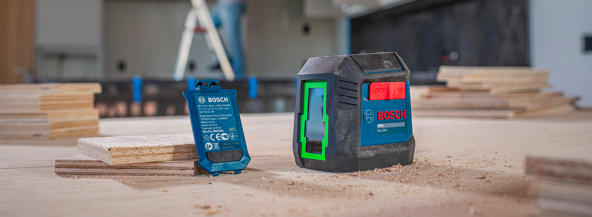 Um nível a laser Bosch Professional está sobre uma superfície de trabalho de madeira, ao lado de uma bateria azul. Pedaços de madeira e serragem estão espalhados, indicando um espaço de trabalho ativo.