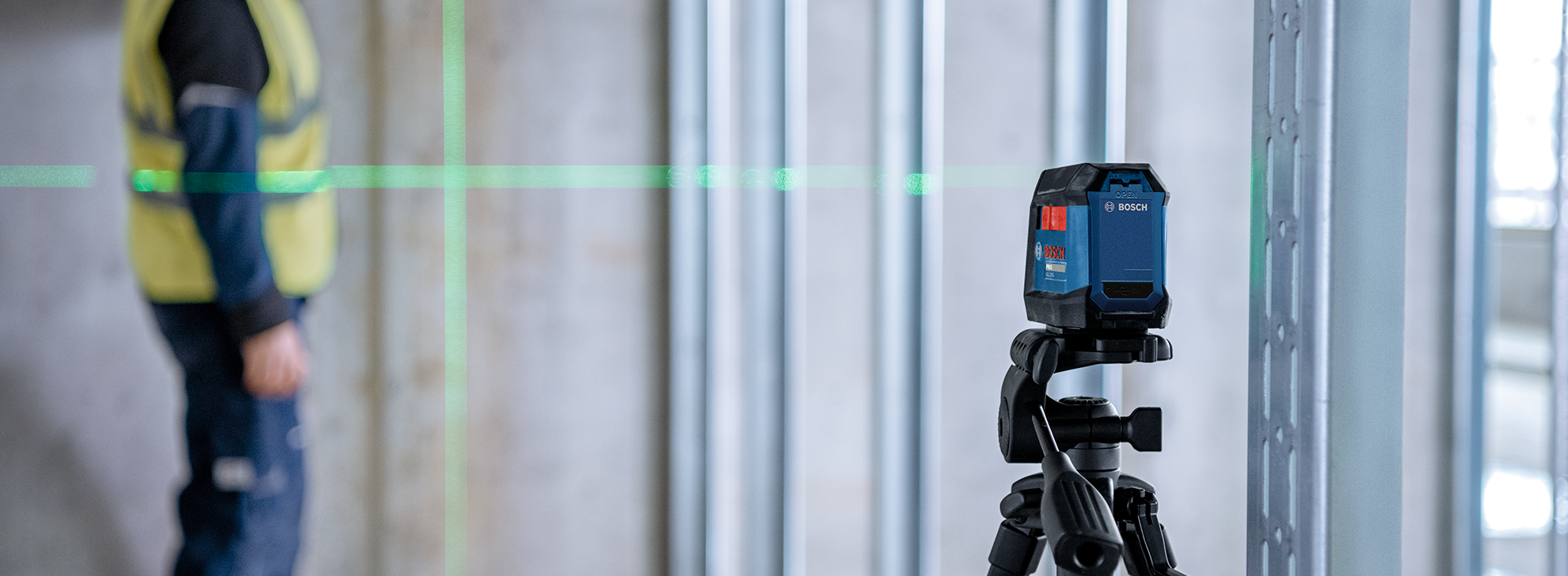 Um nível a laser Bosch Professional em um tripé, com um trabalhador em um colete amarelo desfocado ao fundo.