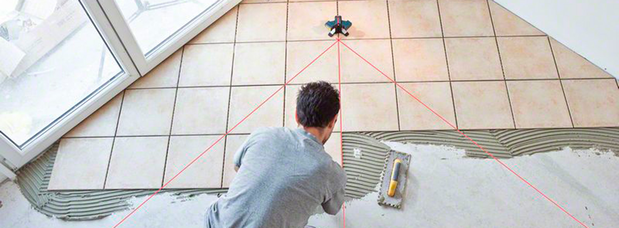 Um homem coloca azulejos no chão usando uma colher de pedreiro e nível a laser.