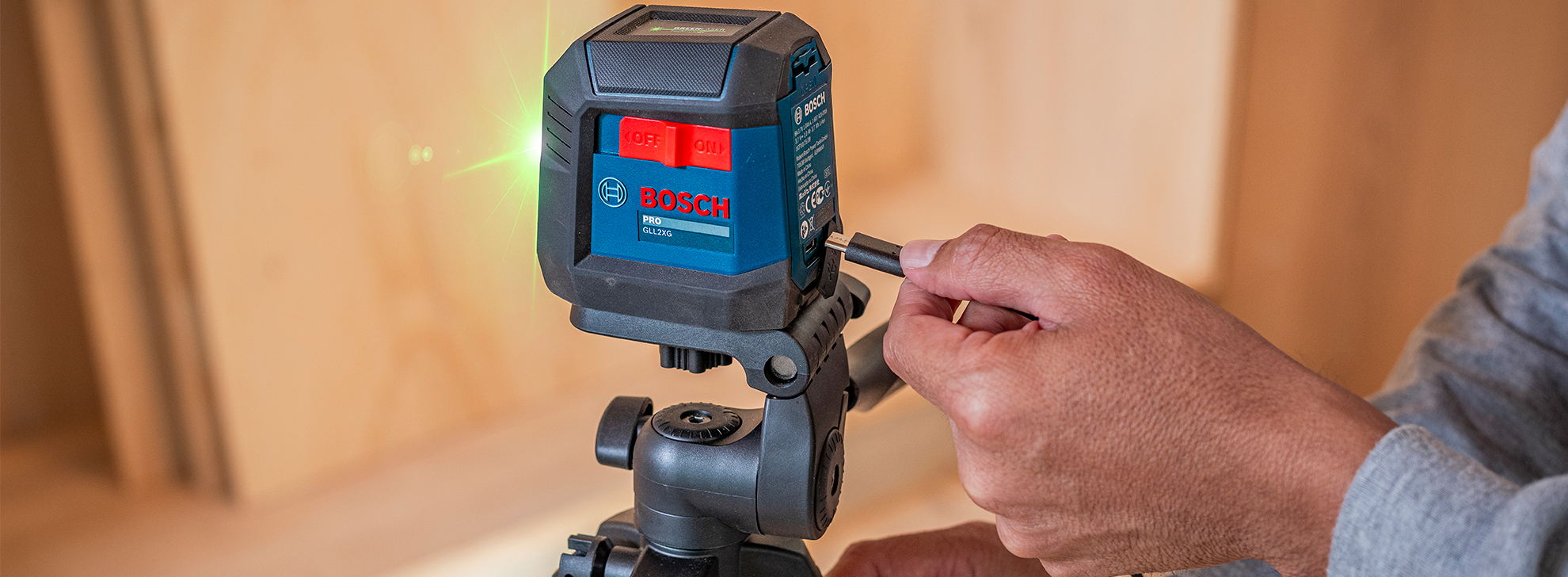 Um nível a laser Bosch Professional montado em um tripé, com uma mão ajustando suas configurações. Feixe de laser verde visível.