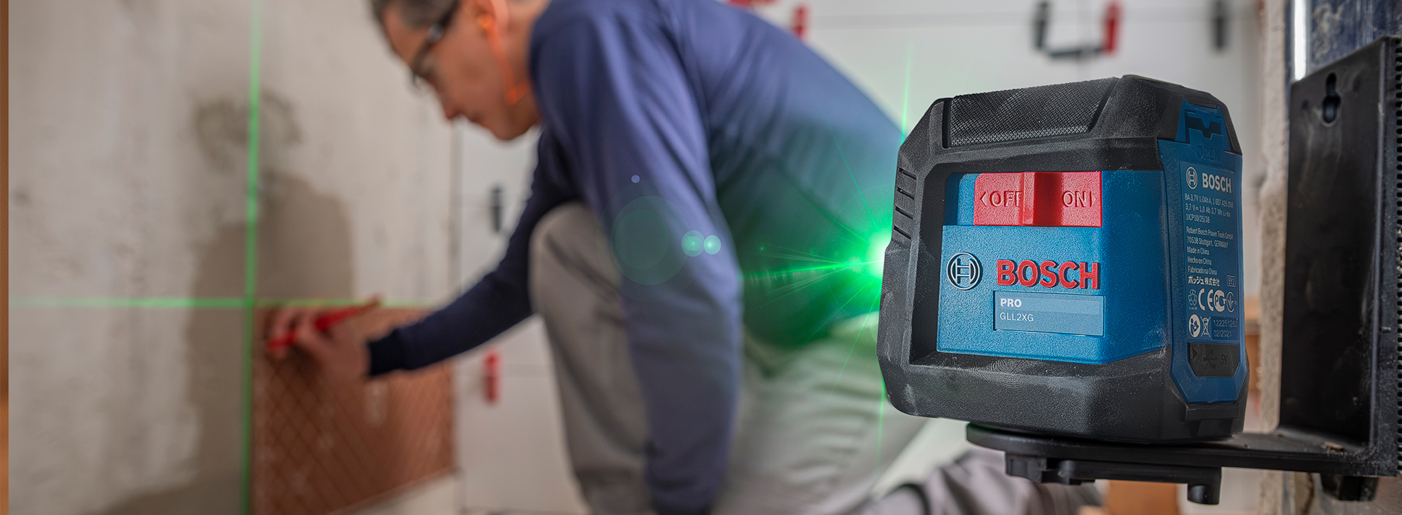 Um nível a laser Bosch Professional está sobre uma superfície, com um trabalhador ajoelhado próximo, alinhando azulejos em uma parede. Linhas de laser verde são visíveis, indicando alinhamento preciso. O trabalhador usa óculos e uma camisa azul, concentrando-se na tarefa.