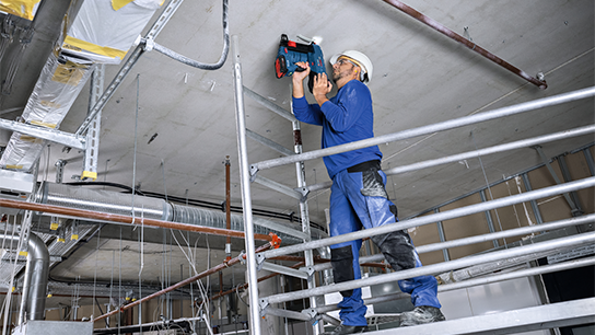Um trabalhador vestido de azul usa uma ferramenta Bosch no teto enquanto está em um andaime.