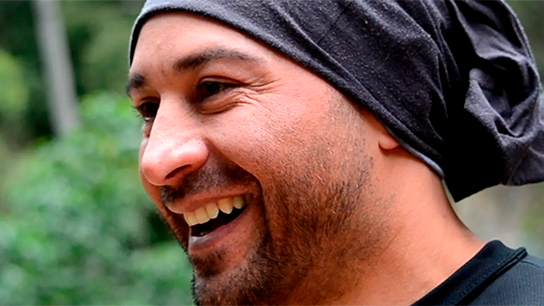 Um homem com uma bandana preta sorri ao ar livre com vegetação ao fundo.