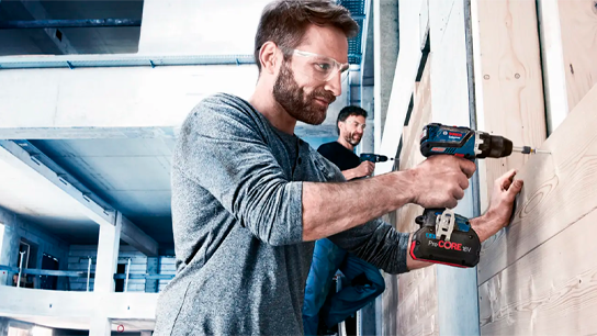 Dois homens trabalham em uma parede de madeira usando furadeiras Bosch em um canteiro de obras.