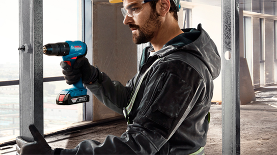 Um homem com uma jaqueta preta usa uma furadeira Bosch azul em uma estrutura metálica.