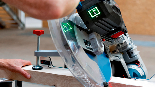 Uma serra de esquadria Bosch Professional corta uma tábua de madeira, com mãos ajustando.