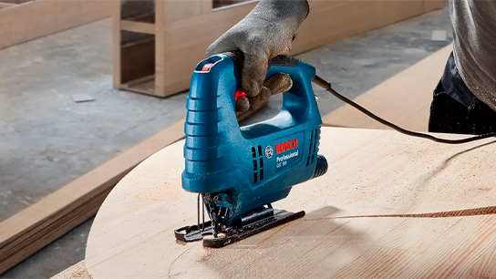 Uma serra tico-tico profissional da Bosch corta madeira, com uma mão segurando-a.