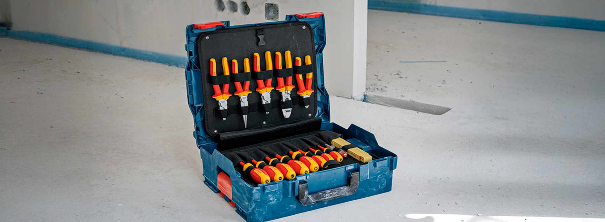 Uma caixa de ferramentas azul da Bosch Professional está aberta em um chão branco, exibindo ferramentas com cabos amarelos dentro.