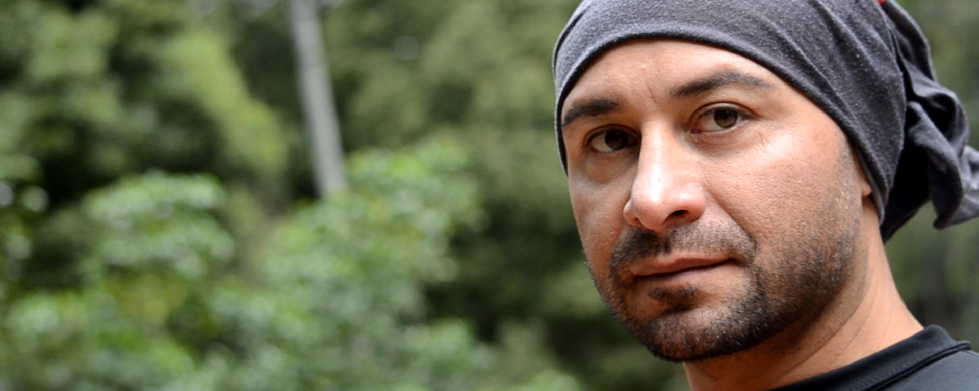 Um homem com uma faixa cinza na cabeça está em um fundo de floresta verde.