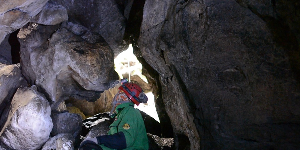 Um espeleólogo com uma camisa verde e capacete explora um túnel de rocha estreito.