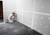 Uma pessoa utiliza uma ferramenta de nivelamento a laser para marcar uma linha reta em uma superfície de drywall.