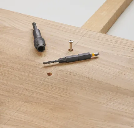 Broca e ponta de chave de fenda sobre uma tábua de madeira ao lado de um parafuso.