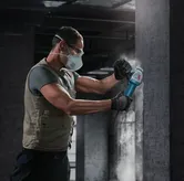 Uma pessoa usando equipamento de segurança usa uma rebarbadora em uma parede de concreto.