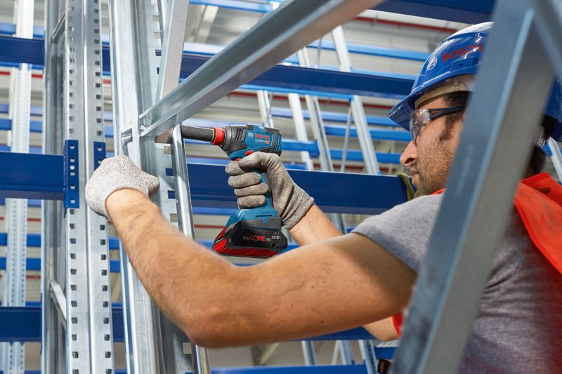 Uma pessoa usando equipamento de segurança usa uma furadeira sem fio em uma estrutura de metal.