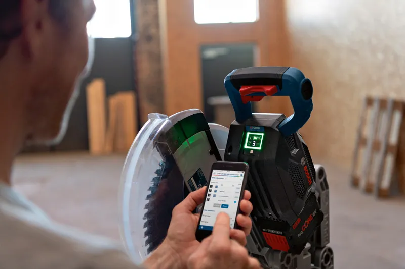 Uma pessoa ajusta uma serra de esquadria sem fio com um smartphone em uma oficina interna.
