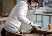 Uma pessoa usando equipamento de segurança lixa uma perna de mesa de madeira em uma oficina.