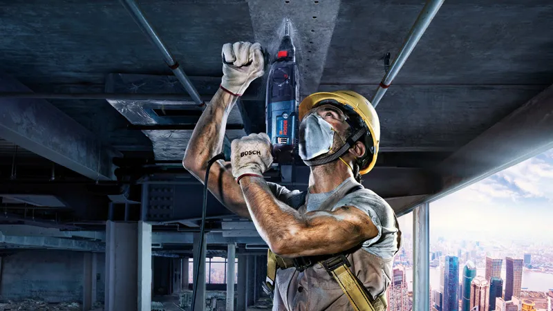 Um trabalhador usando equipamento de segurança perfura um teto de concreto em um canteiro de obras.