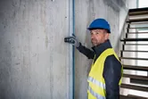 Uma pessoa usando equipamento de segurança utiliza um medidor a laser contra uma parede de concreto.