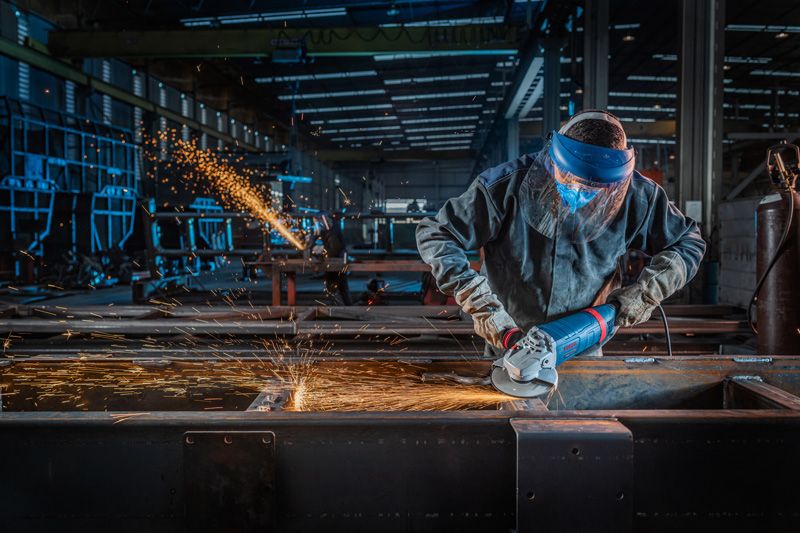 Uma pessoa usando equipamento de segurança tritura metal com uma esmerilhadeira angular, produzindo faíscas.