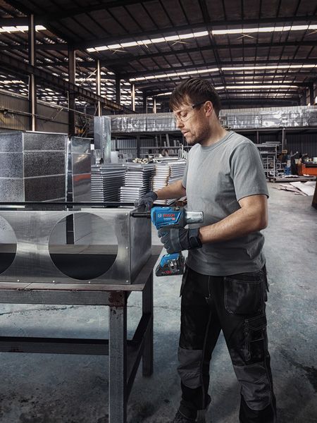 Uma pessoa usando equipamento de segurança faz furos em dutos de ar de metal em uma fábrica.