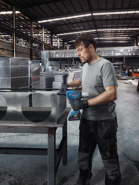 Uma pessoa usando equipamento de segurança faz furos em dutos de ar de metal em uma fábrica.