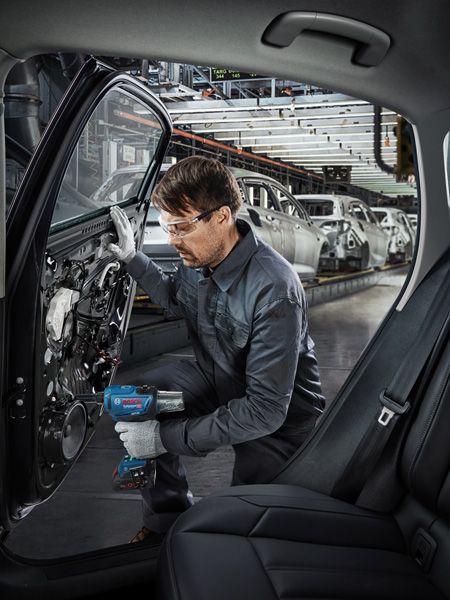 Uma pessoa usando equipamento de segurança usa uma ferramenta elétrica para montar uma porta de carro em uma fábrica automotiva.