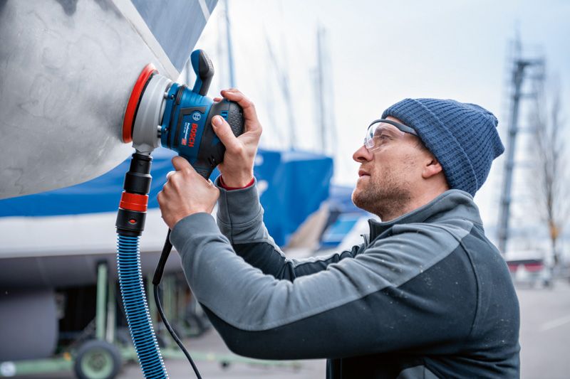 Uma pessoa usando equipamento de segurança lixa o casco de um barco com uma lixadeira orbital aleatória.