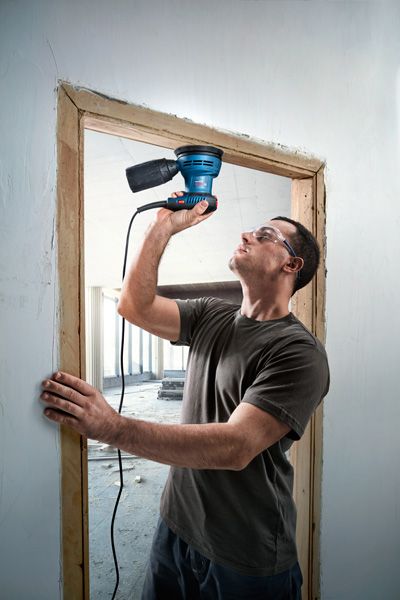 Uma pessoa usando equipamento de segurança lixa uma moldura de porta de madeira com uma lixadeira elétrica.