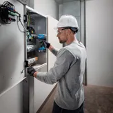 Uma pessoa usando equipamento de segurança usa uma furadeira sem fio em um painel elétrico.