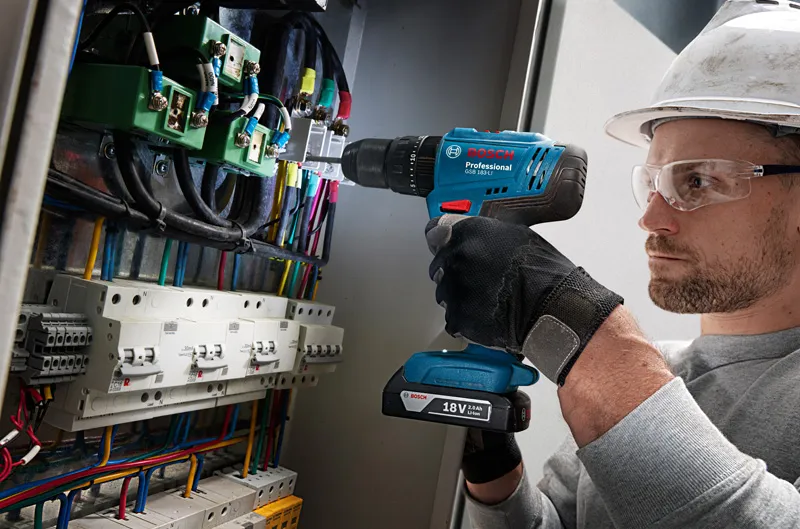Uma pessoa usando equipamento de segurança perfura um painel de controle cheio de fios.